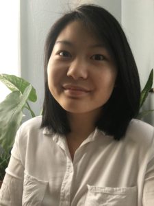 A photo of Macayla smiling at the camera, wearing a white button up shirt, in front of some plants.